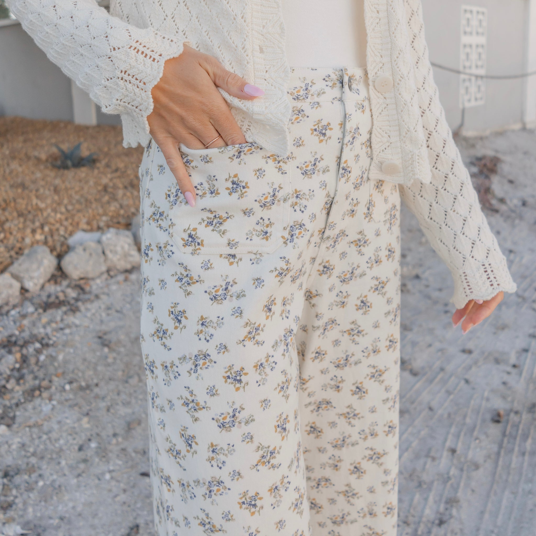 Person wearing a white lace cardigan and floral pants standing on a pebbled surface.
