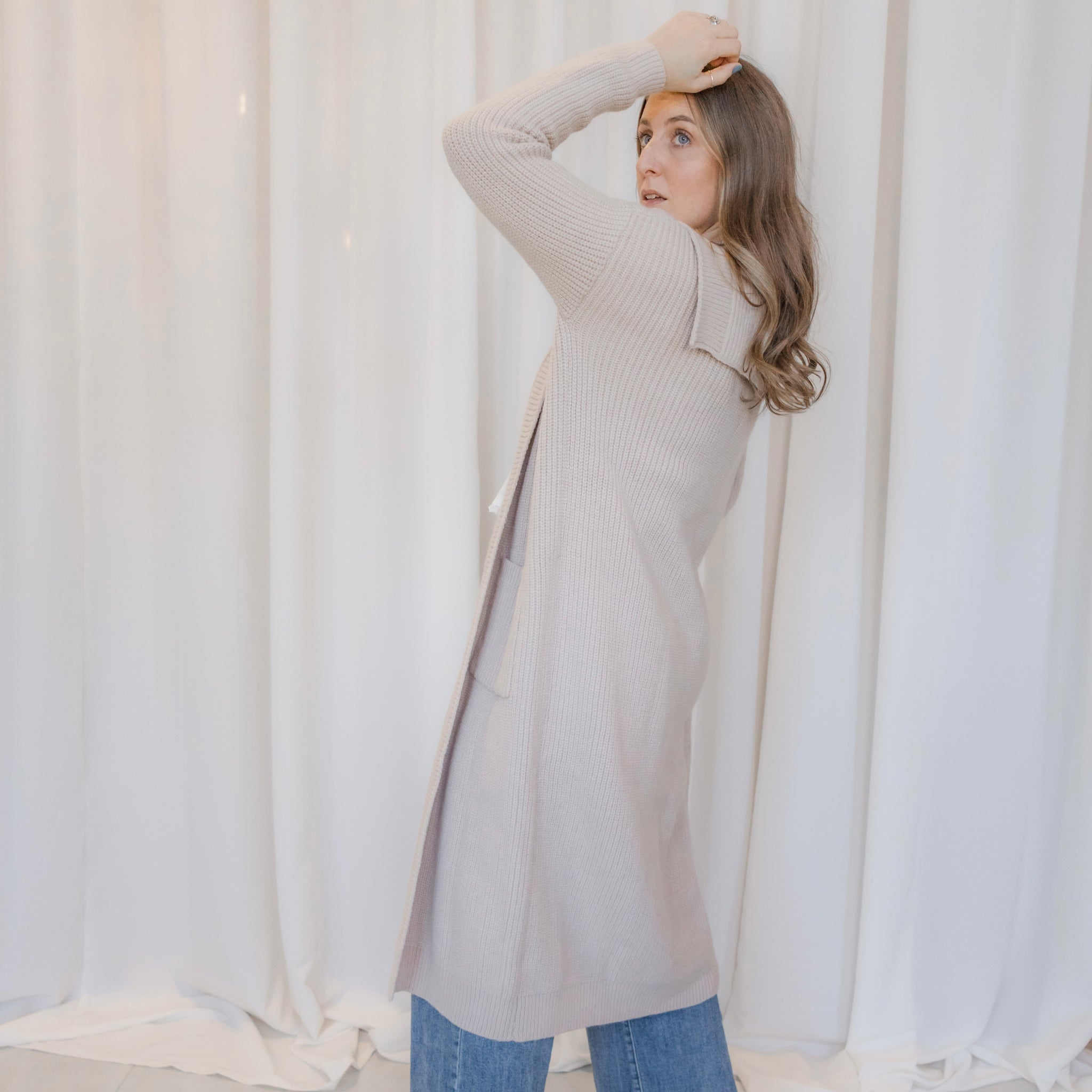 Woman wearing a long beige cardigan in front of white curtains