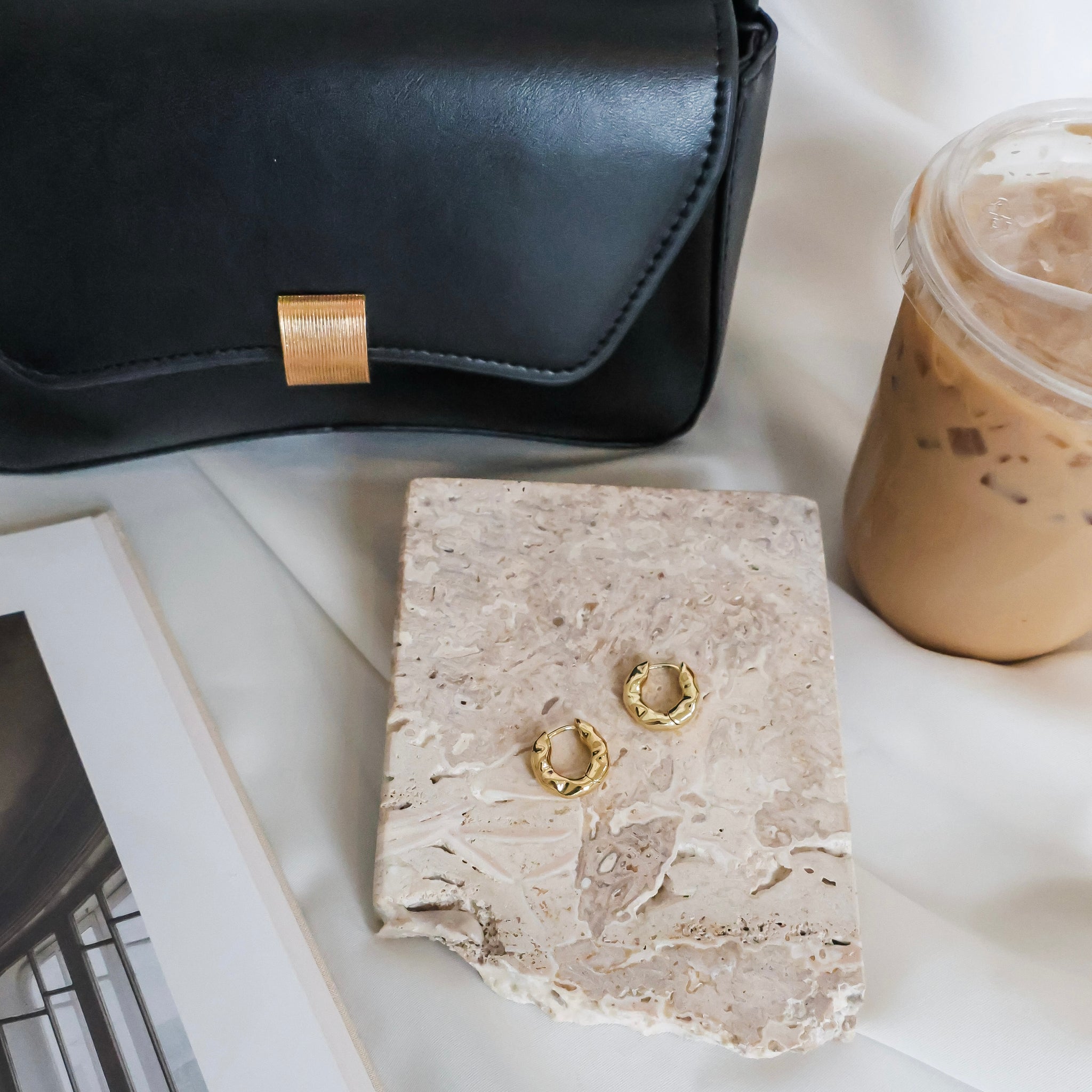Gold hoop earrings on a stone surface with a black handbag and iced coffee in the background.
