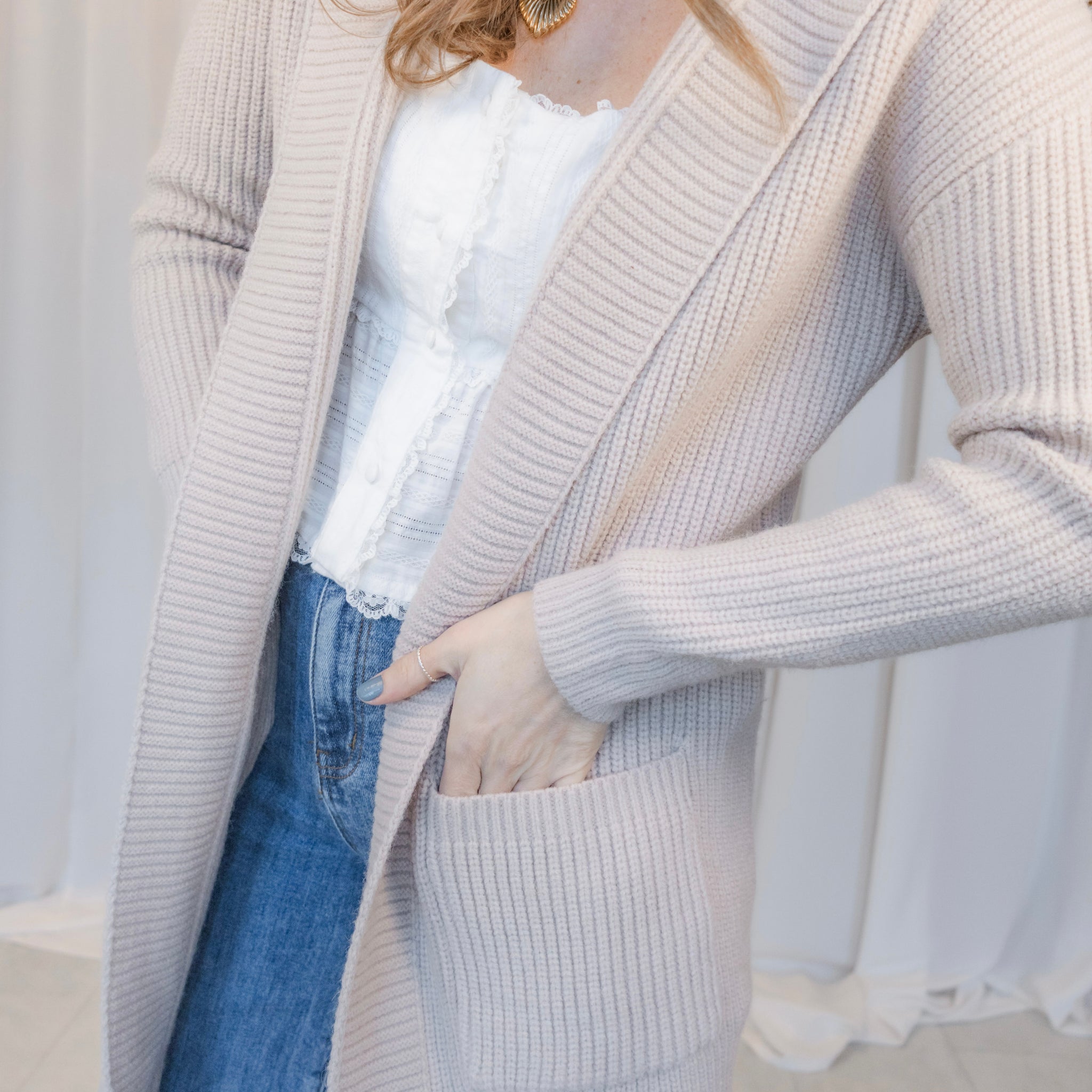 Person wearing a long beige cardigan over a white top and blue jeans, standing against a neutral background.
