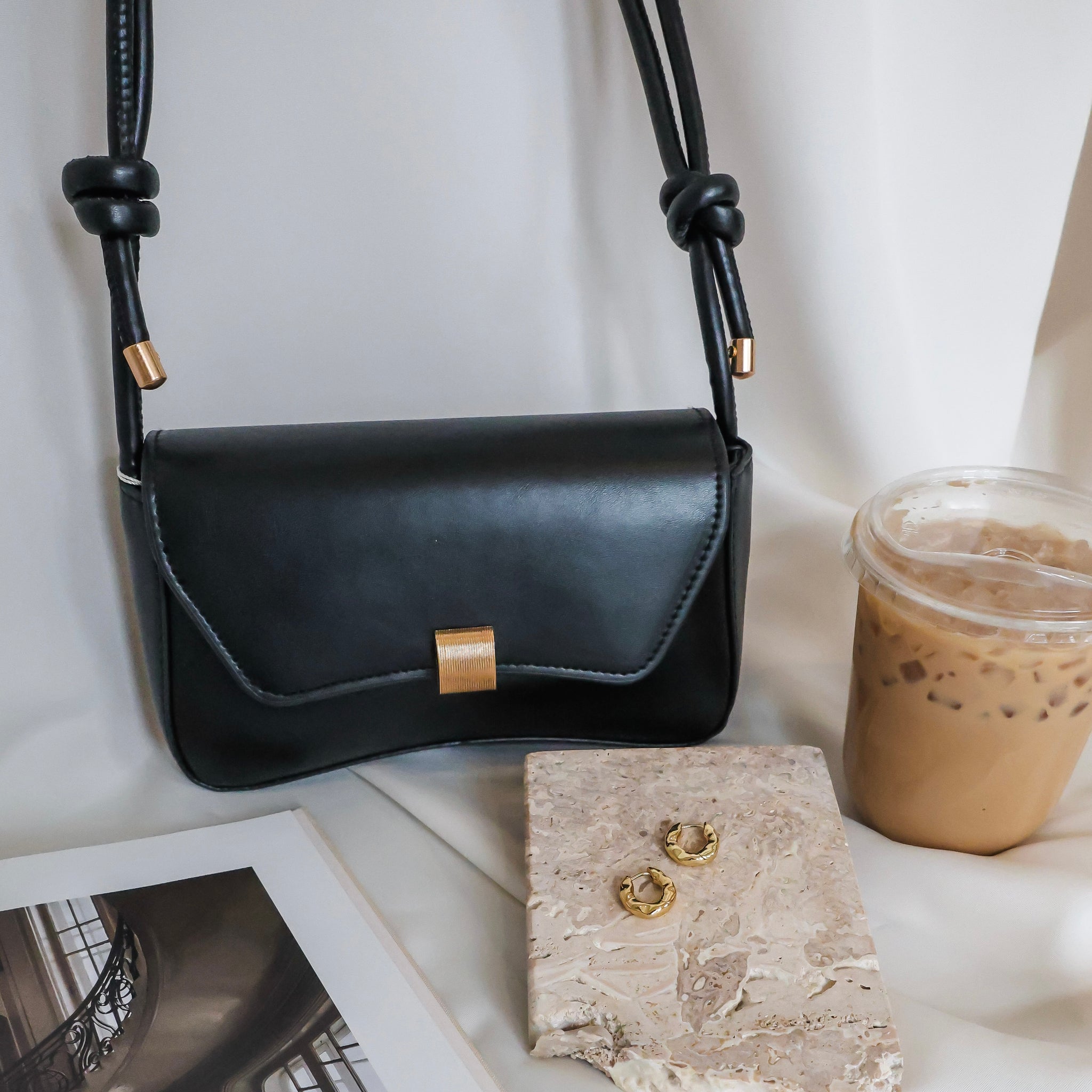 Black leather handbag with gold accents on a white surface with a drink and jewelry.