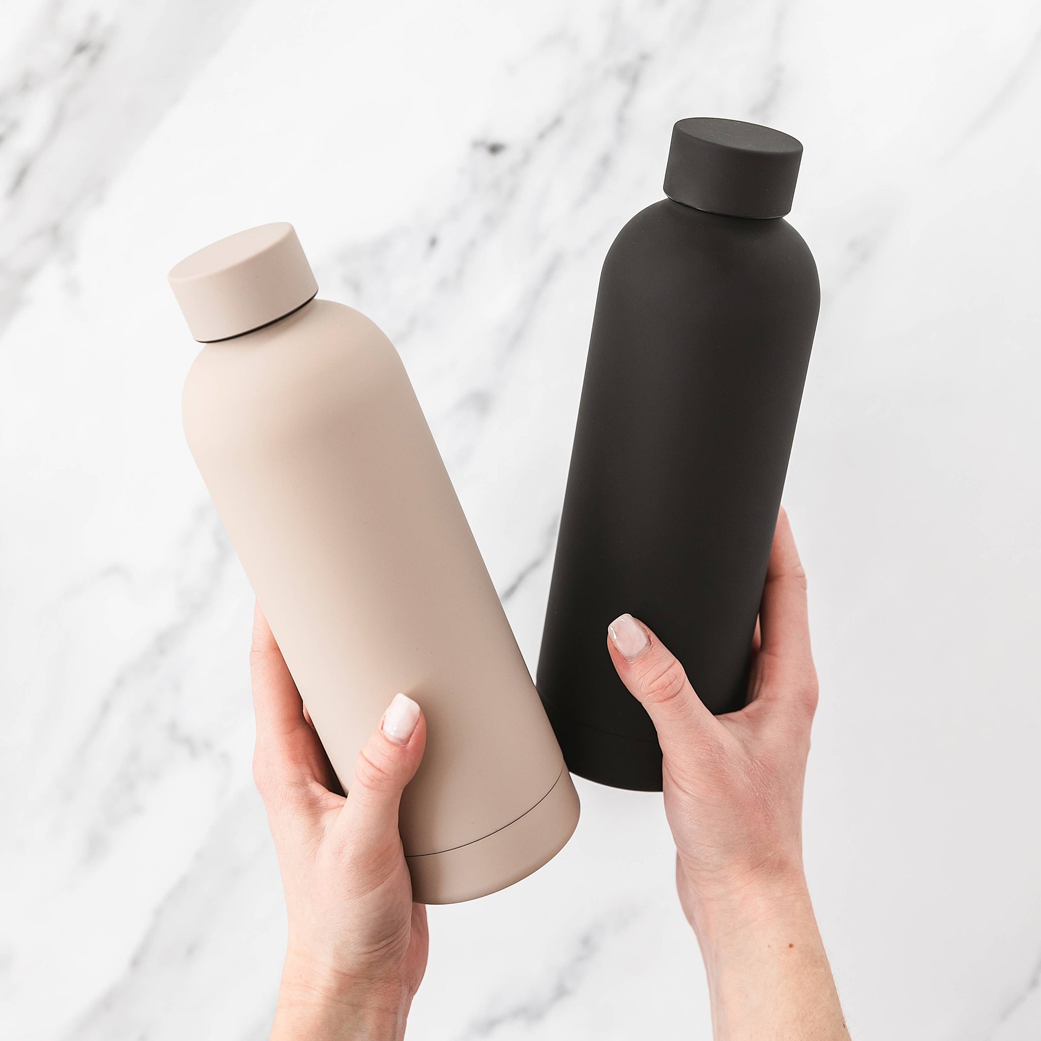 Two water bottles, one beige and one black, held by a person against a light background.