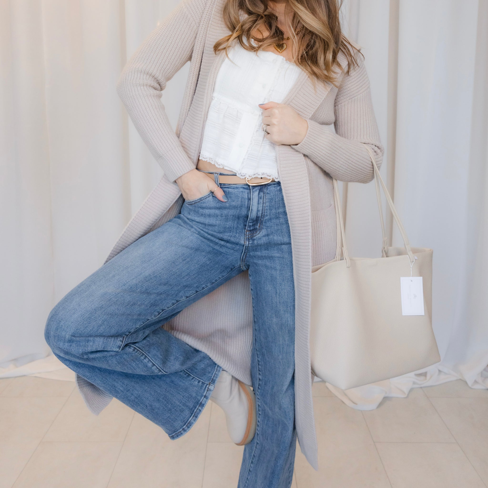 Woman wearing a beige cardigan, white shirt, and blue jeans, holding a beige tote bag.