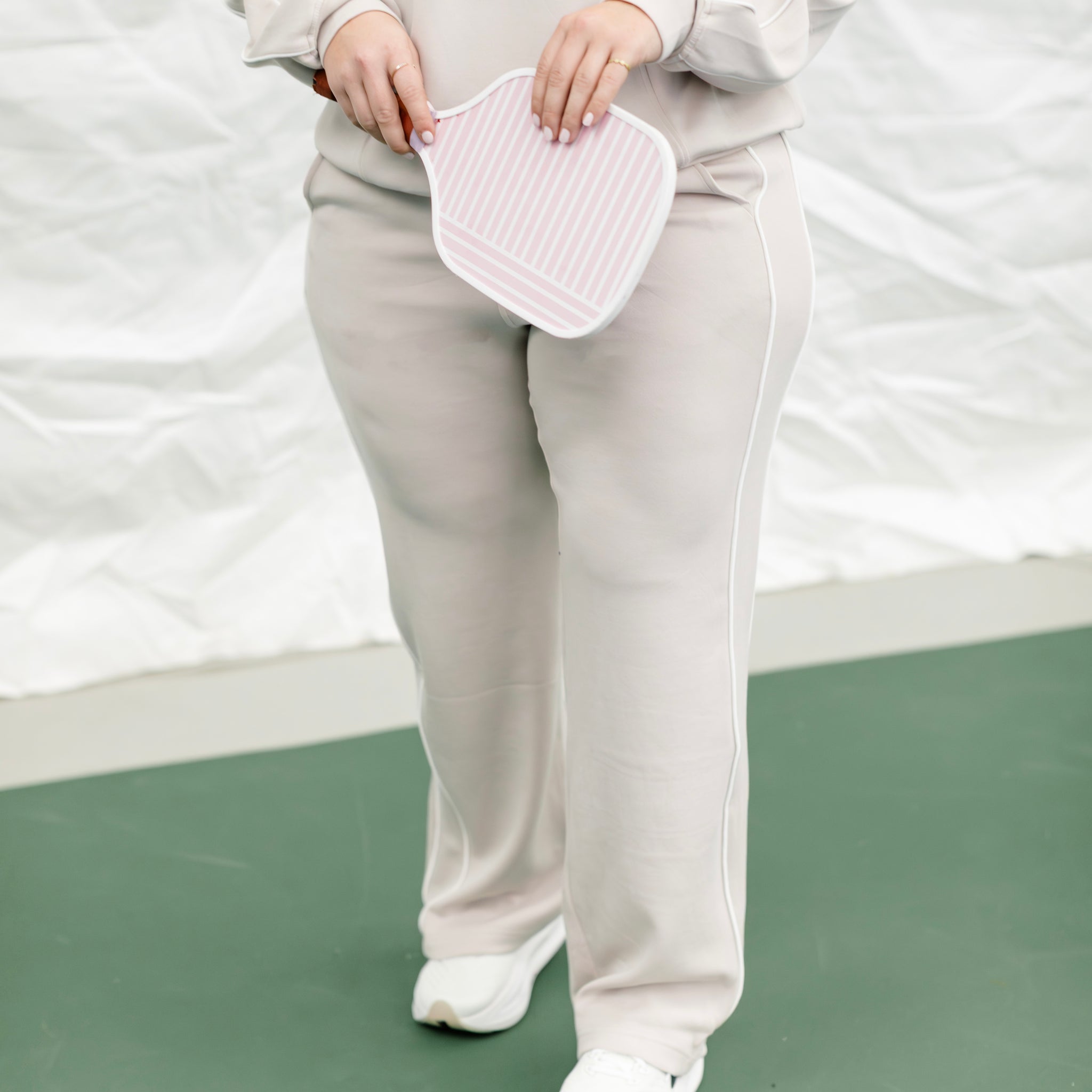Woman wearing a beige track suit with white piping on the side while holding a pickleball paddle. 