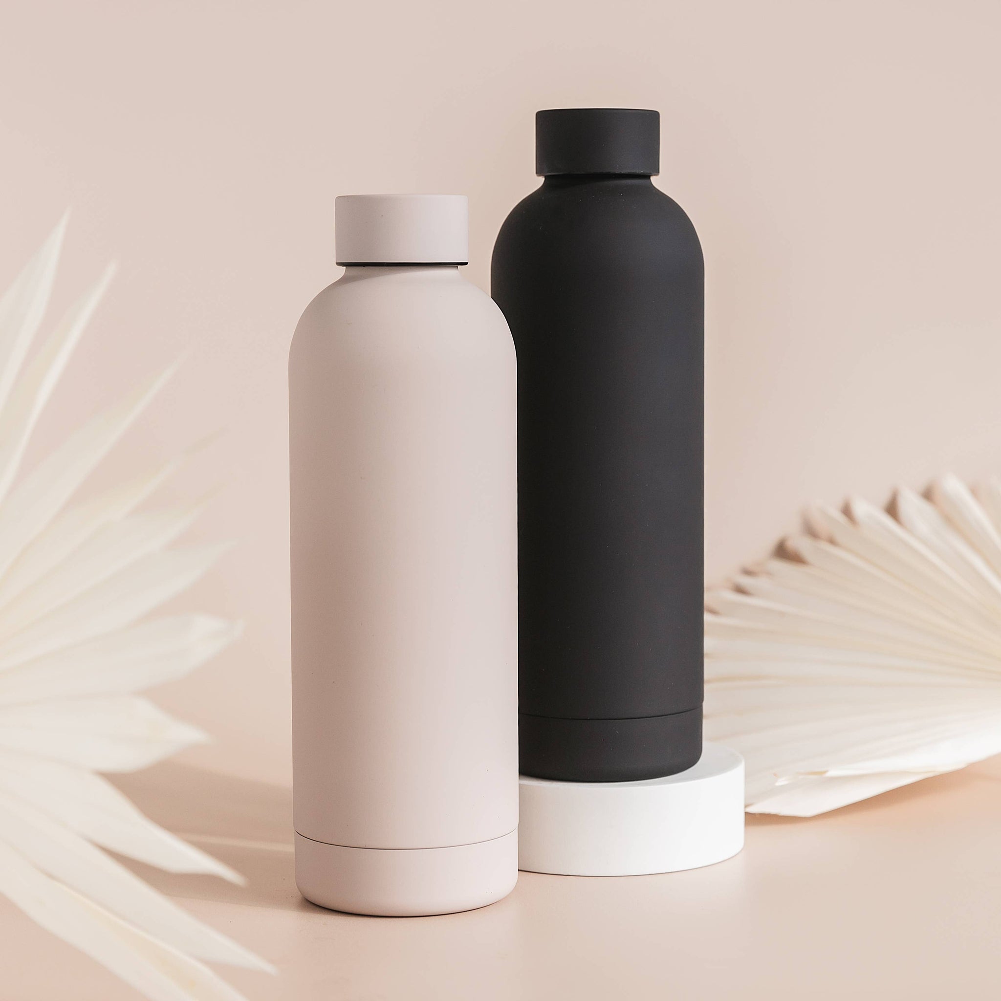Two insulated water bottles, one black and one beige, on a light pink background with leafy textures.