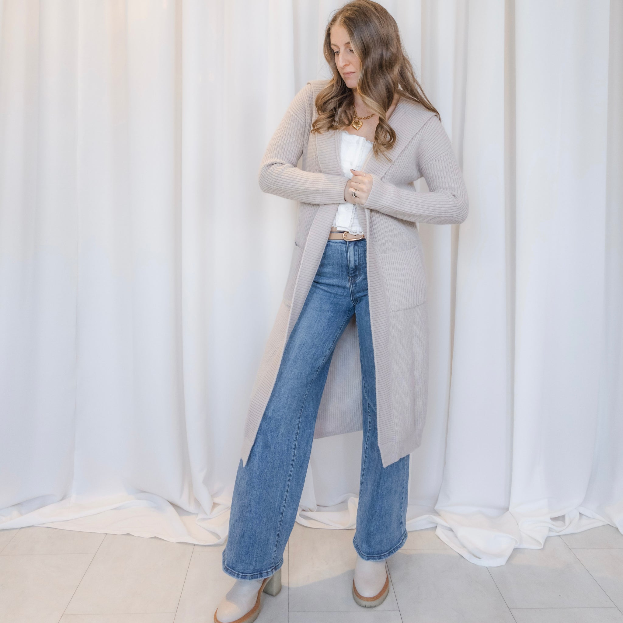 Woman wearing a long gray cardigan, white top, and blue jeans against a white curtain background.