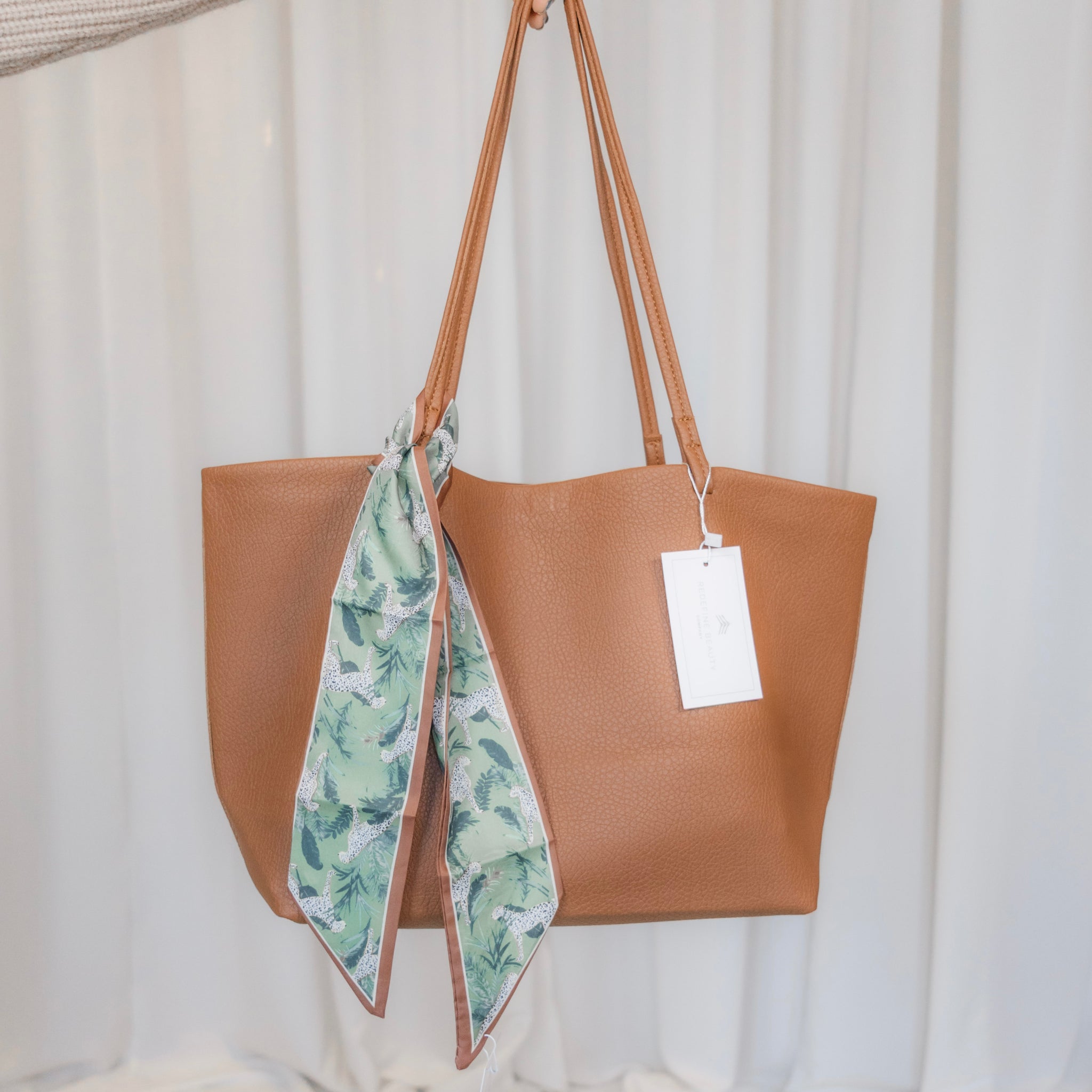 Brown tote bag with a green leaf-patterned scarf draped over it against a white background