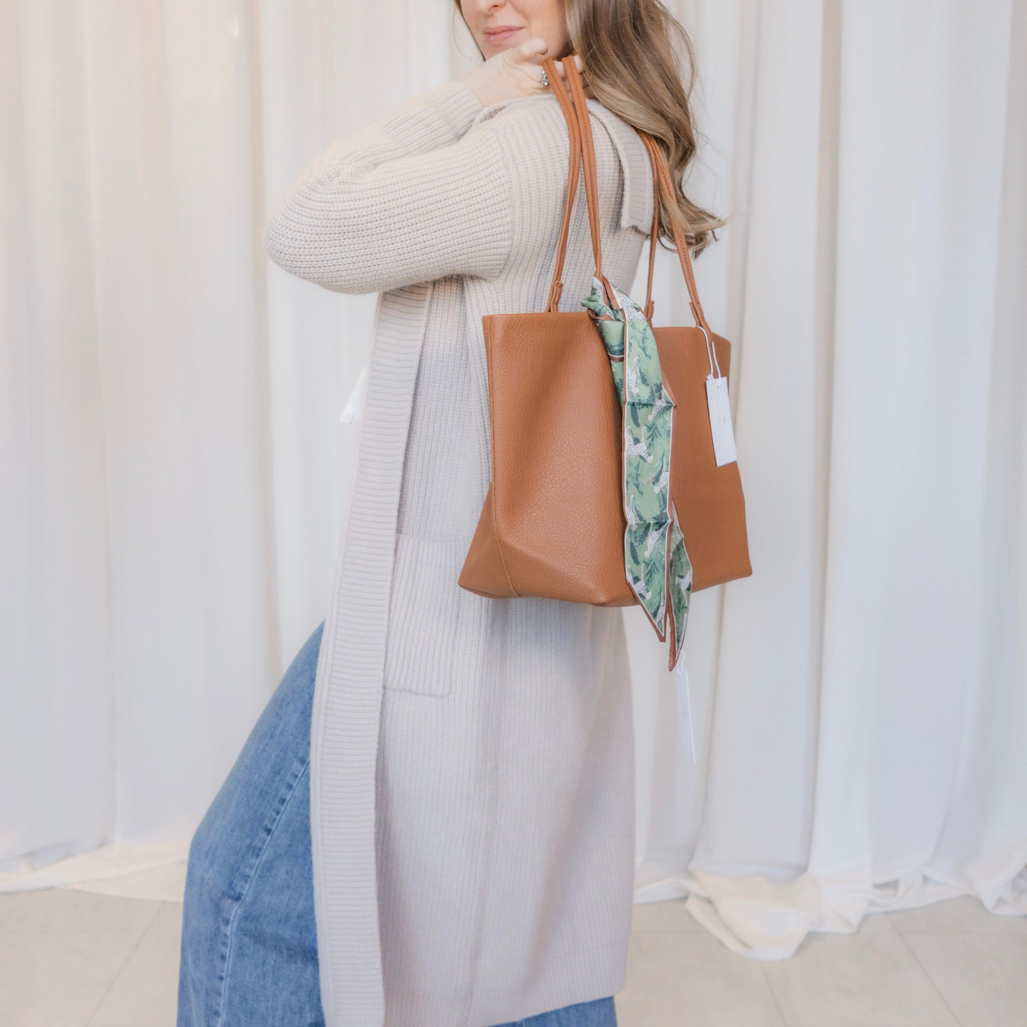Woman holding a brown tote bag with a scarf inside against a white curtain background