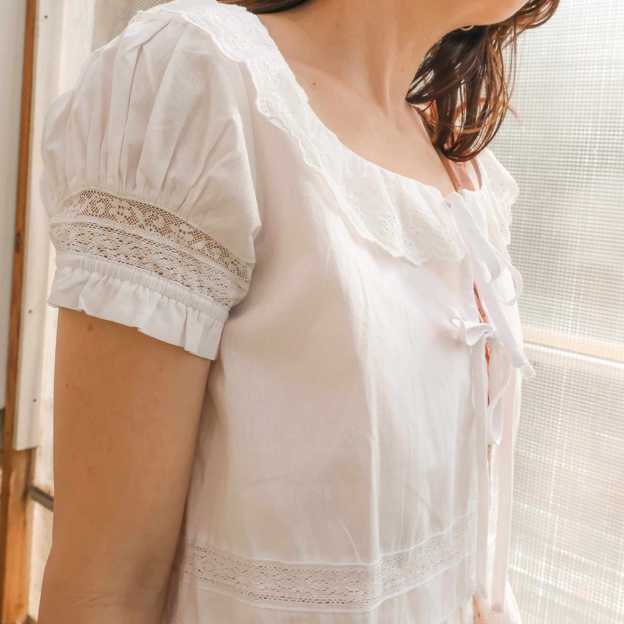 Woman wearing a white blouse with lace details in a softly lit room.