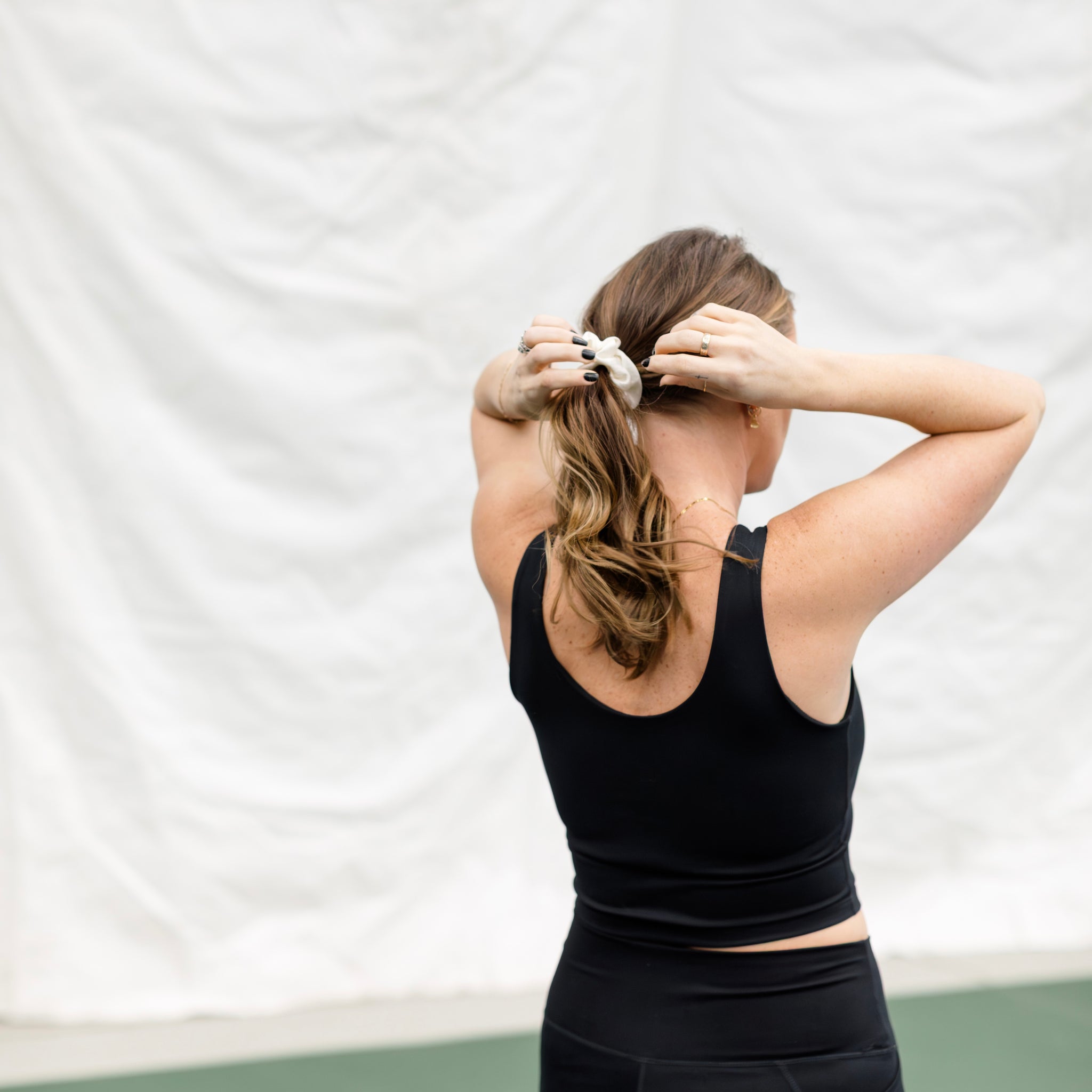 Woman wearing a black athletic set on a pickleball court. Showing a view of the back and fixing her ponytail. 