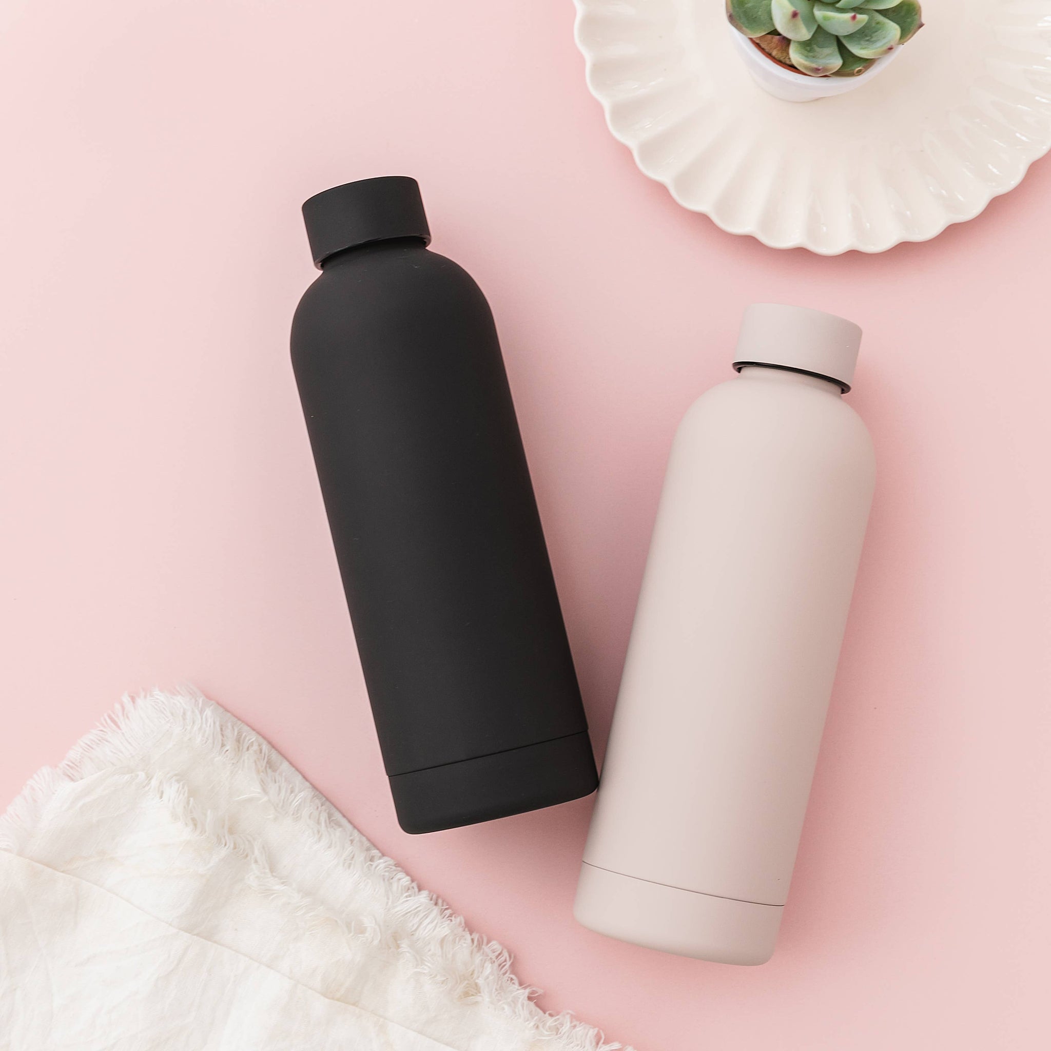 Black and beige water bottles on a pink surface with a white cloth and plant.