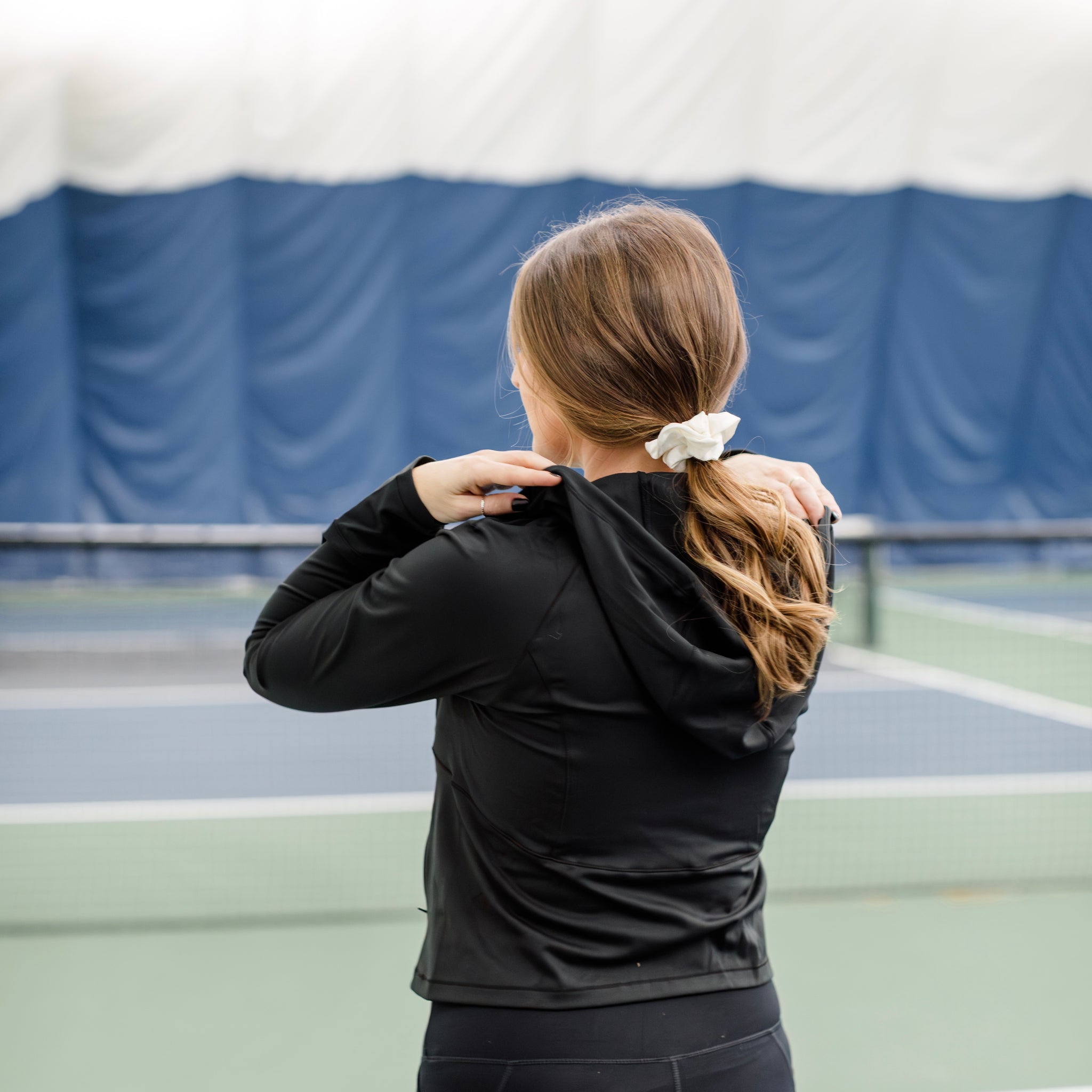 Woman wearing a track jacket on a pickleball court while showing the back and hood details. 