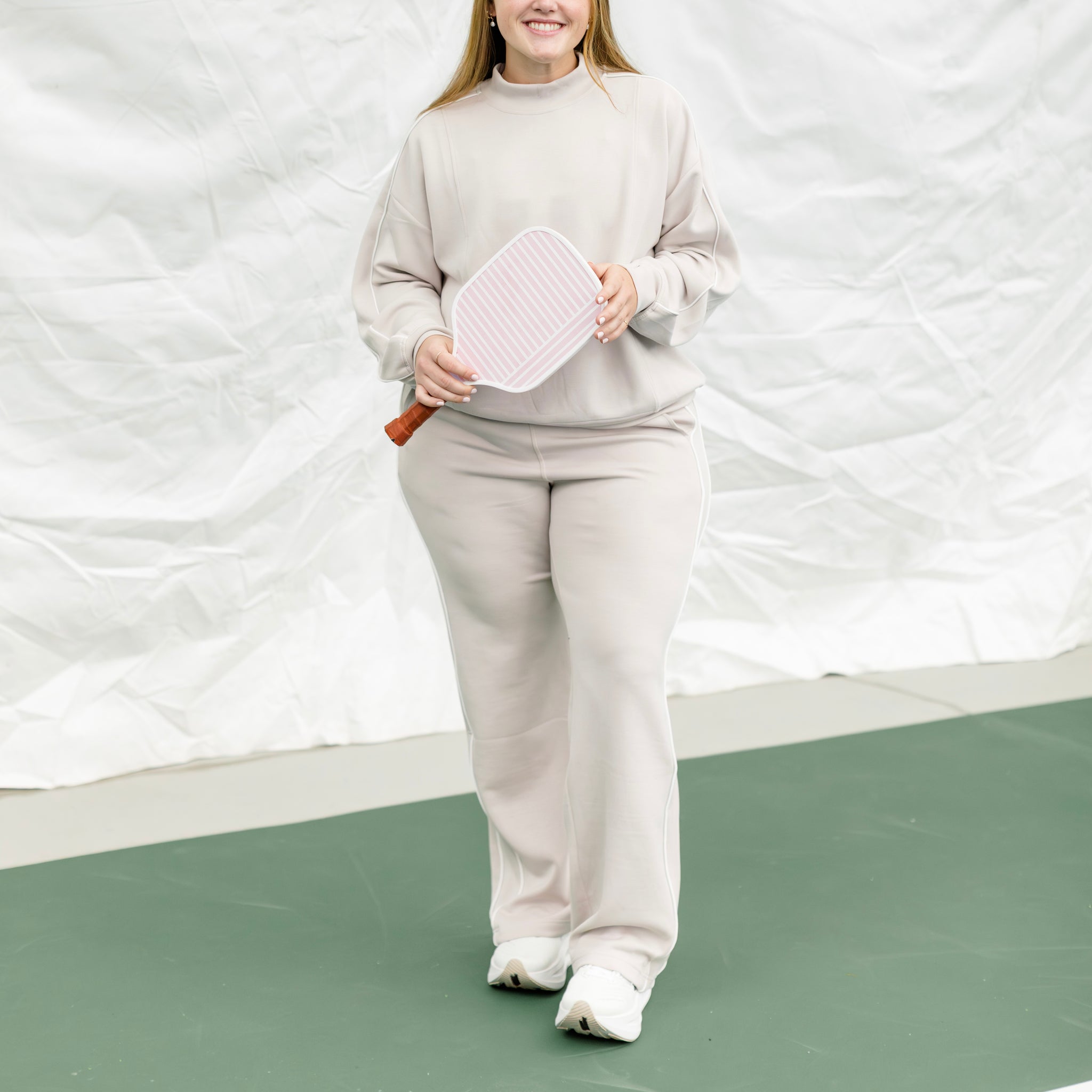 Woman wearing a beige track suit with white piping on the side, holding a pickleball paddle.