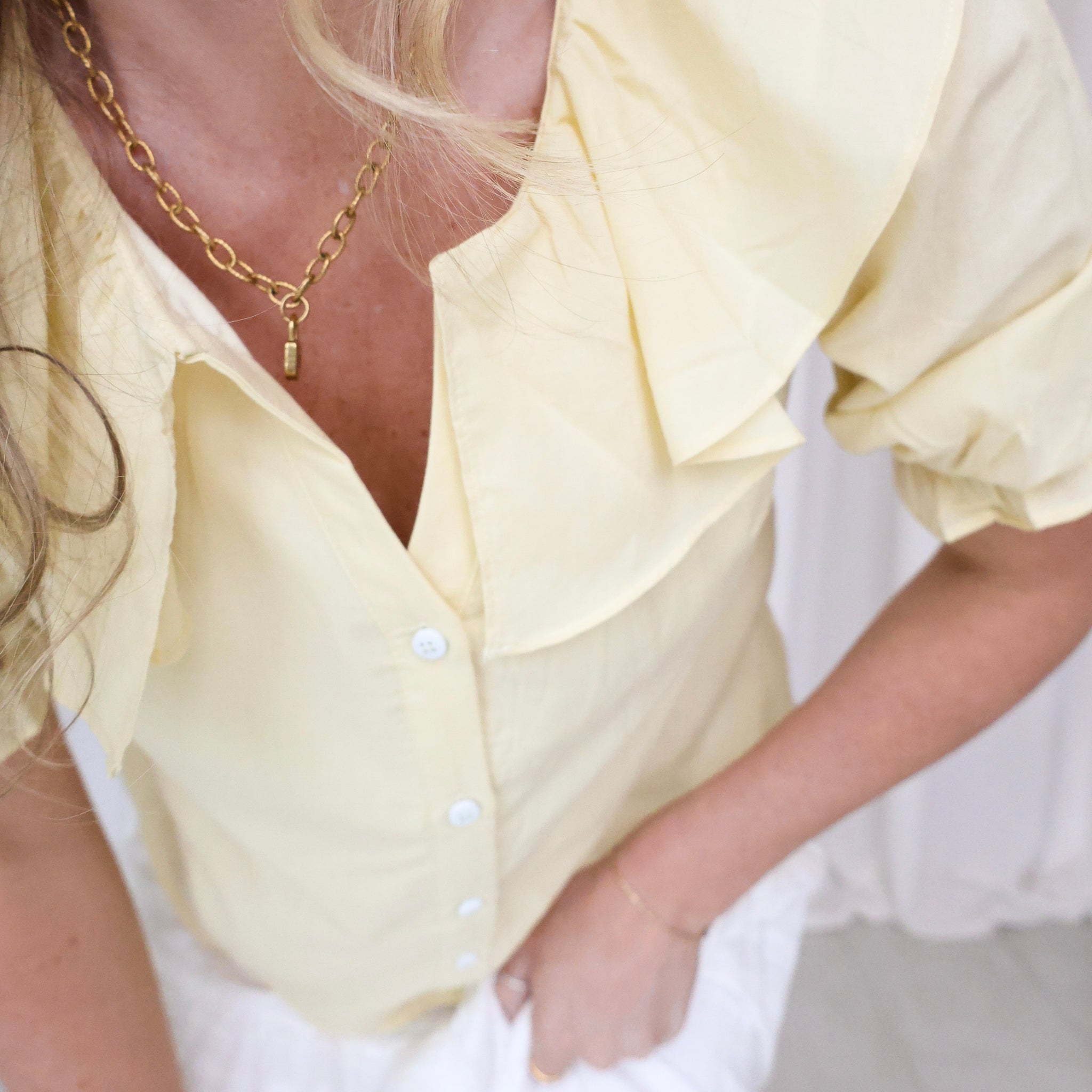 Person wearing a light yellow blouse with ruffled sleeves and a gold necklace.