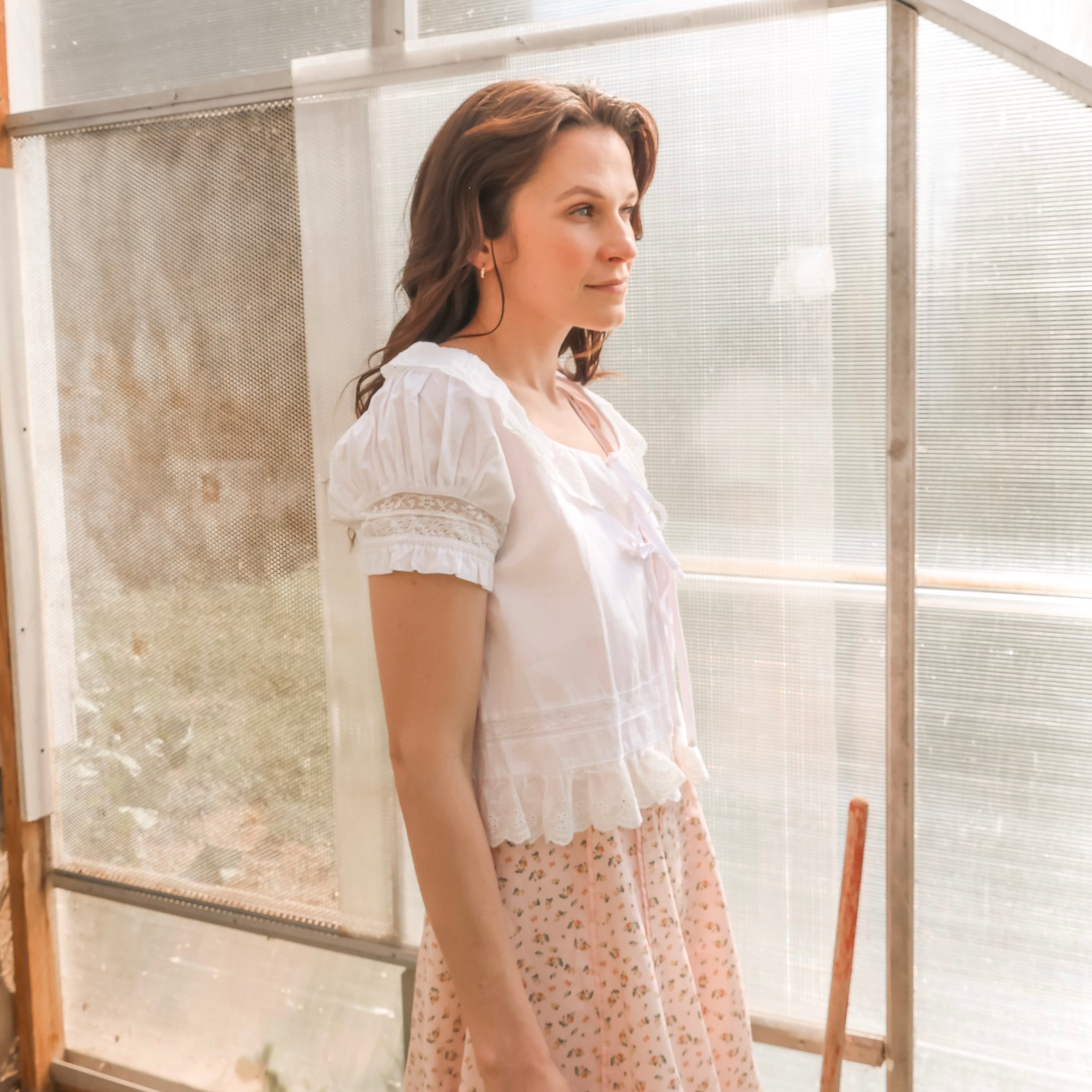 Woman in a white blouse and floral skirt standing in a greenhouse.