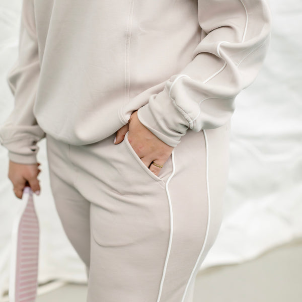 Woman wearing a beige track suit with white piping on the side, she has her hand in her pocket. 