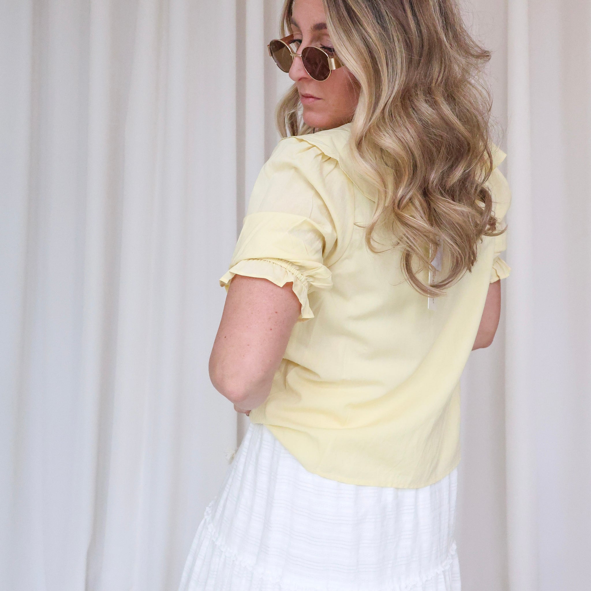 Woman wearing a light yellow blouse with ruffled sleeves and a white skirt against a plain background