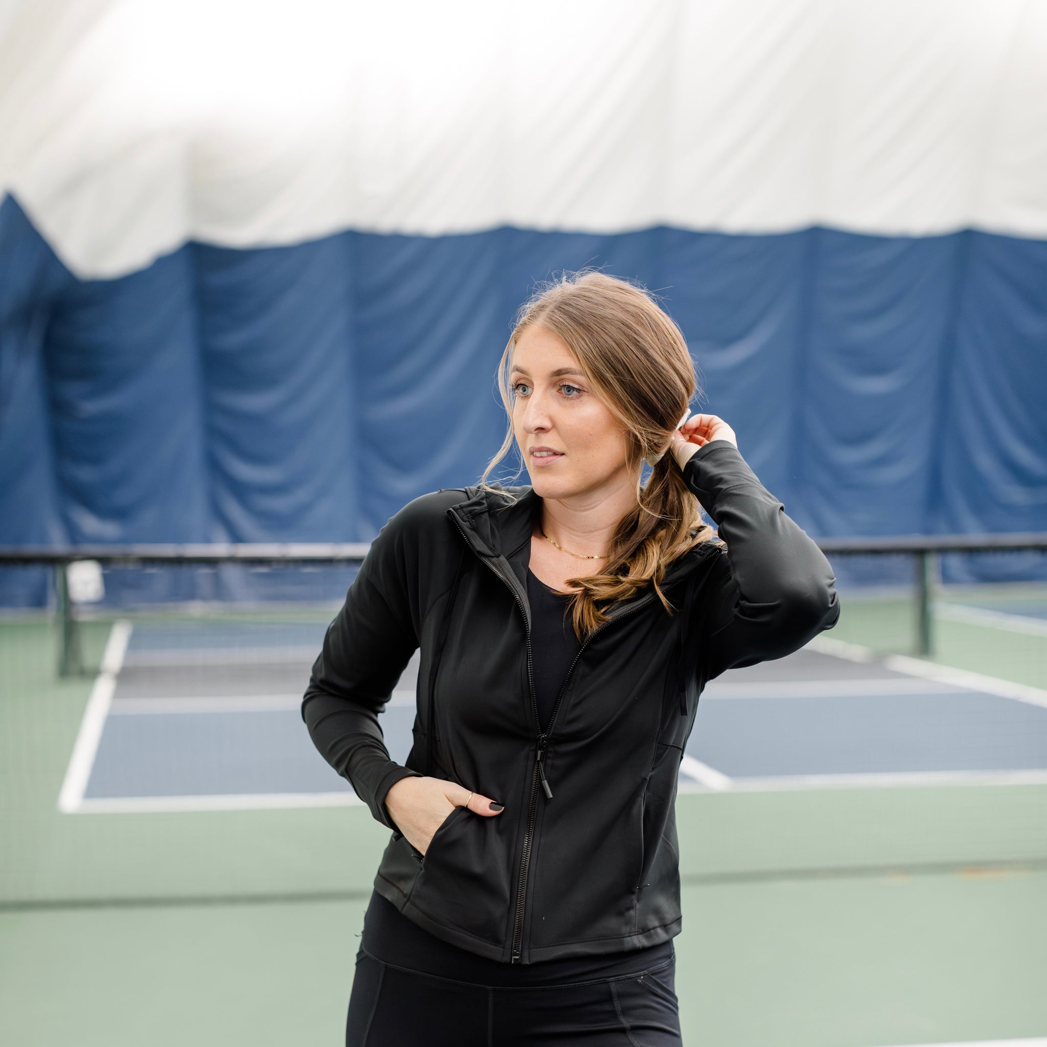 Woman wearing a track jacket on a pickleball court with one hand in her hair. 